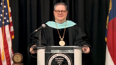 Darrin Hartness at lecturn between American and North Carolina flags in graduation robes.