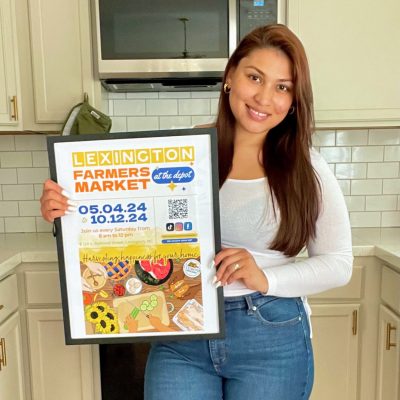 Veronica Orellana holding winning Farmer's Market poster design. Poster text reads: "Lexington Farmers Market at the Depot 05.04.24 to 10.12.24. Join us every Saturday from 8 a.m. to 12 p.m. Harvesting happiness for your home."
