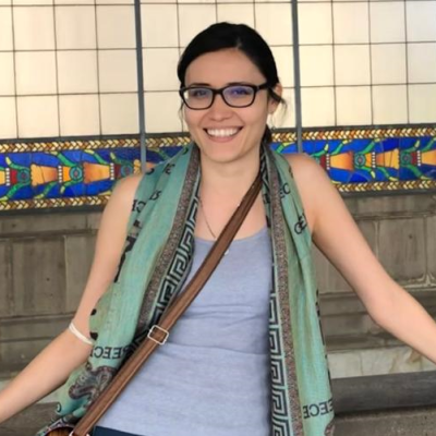 Smiling woman in glasses with a scarf and a crossbody bag, standing in front of a tiled wall with a colorful mosaic pattern.