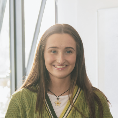Smiling young woman with long brown hair, wearing a green sweater and a flower pendant necklace, seen indoors with large windows in the background.
