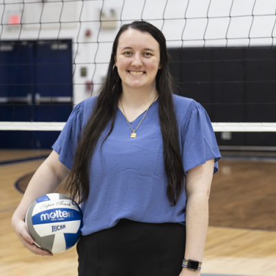 Woman with long dark hair holding a Molten volleyball in a gym with a volleyball net in the background.