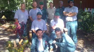 A group of nine men outdoors in a garden, wearing gray and green clothing, surrounded by foliage and a display of vegetables.