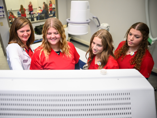 Four women in a medical training room view a computer screen; one in a white coat smiles while pointing.