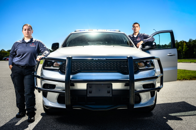 Two uniformed individuals standing beside a white law enforcement vehicle with a grille guard.