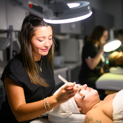 A woman in a spa applies a facial mask to a client's face under a circular lamp.