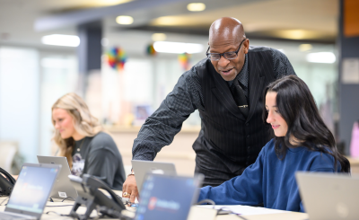A man in a black vest assists a woman at a laptop in a modern, bright learning environment.