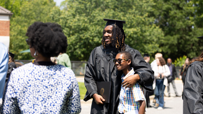 Graduate in cap and gown with young boy at an outdoor ceremony.