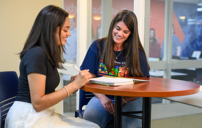 Two people sitting at a table, one writing in a notebook and both smiling, with a glass wall in the background.