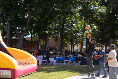 People playing basketball at an outdoor community event with an inflatable hoop game and tables set up under trees.
