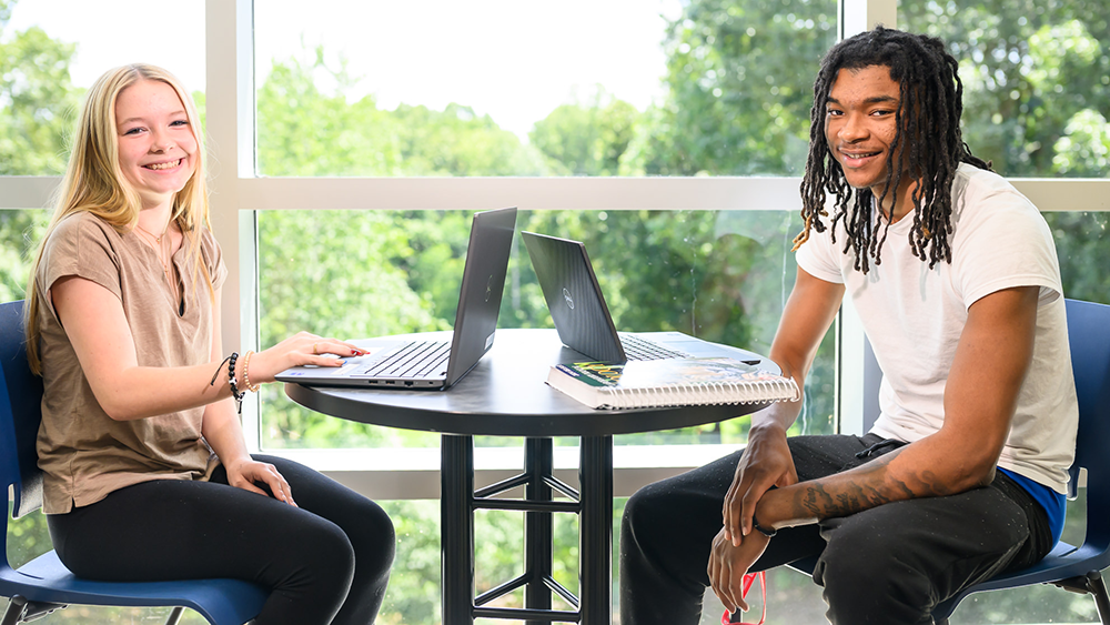 Two people sitting at a table with laptops in a bright room next to large windows.
