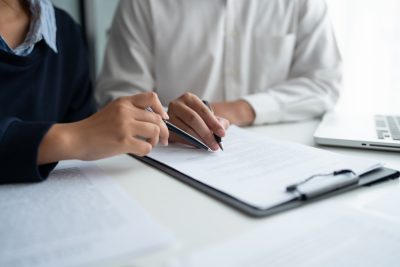 Two people reviewing documents at a desk with a clipboard and laptop.