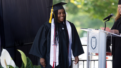A graduate in a cap and gown smiles on stage during a graduation ceremony next to a podium.