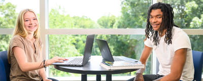 Two people sitting at a table with laptops in a bright room next to large windows.