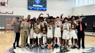 A jubilant basketball team celebrates with coaches on a gym court, holding championship plaques.