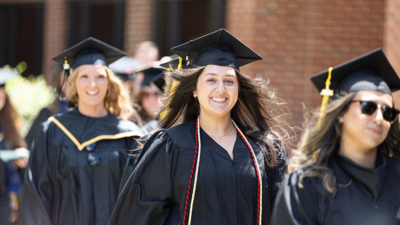 A group of graduates in black caps and gowns walk outdoors during a commencement procession, smiling as they head toward the ceremony.