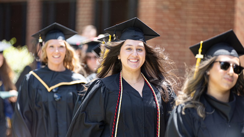 A group of graduates in black caps and gowns walk outdoors during a commencement procession, smiling as they head toward the ceremony.