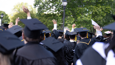 Graduates in black gowns and caps raise their arms outdoors under green trees.
