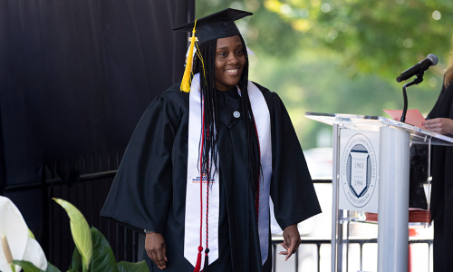 A graduate in a cap and gown smiles on stage during a graduation ceremony next to a podium.